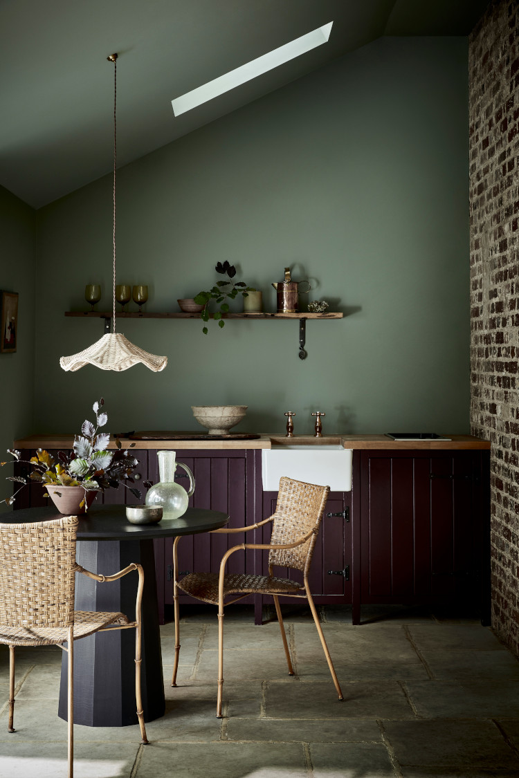 A olive green coloured kitchen with flagstone tile floor, dark brown cabintery which surrounds a small 2 person black table