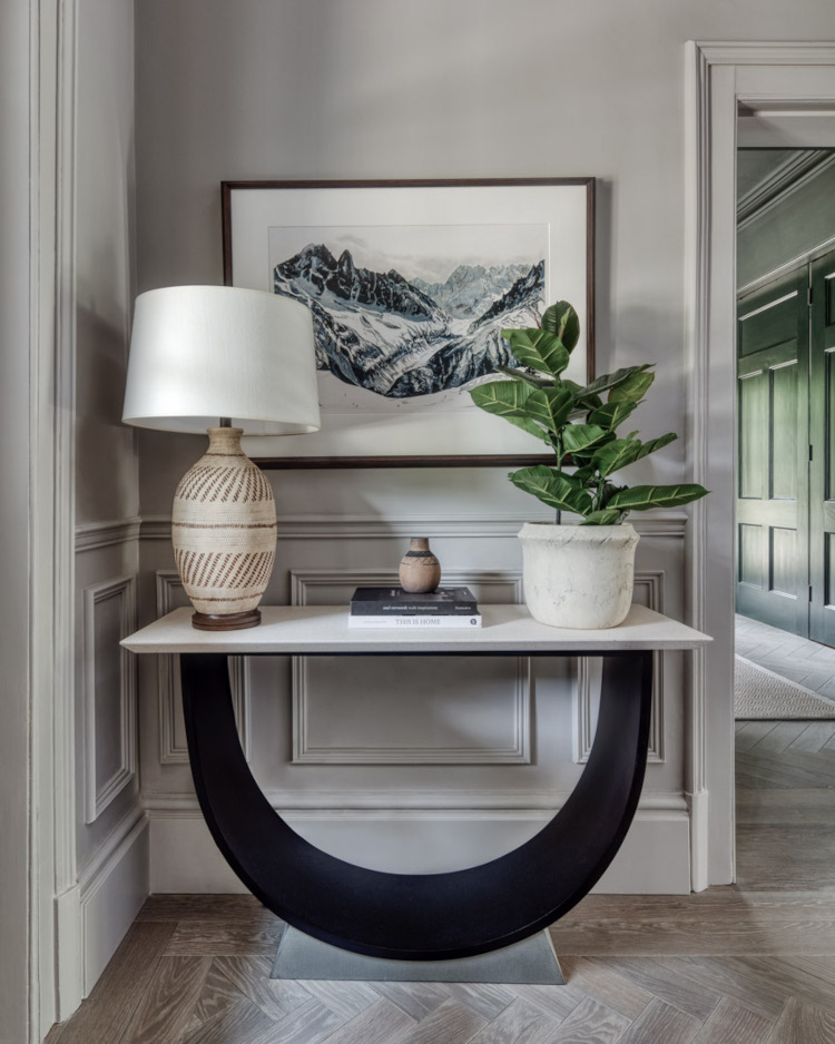A large wood panelled hallway houses a large U shaped dark wood table on which sits a large ceramic lamp and plant. Behind is a large linoprint artwork showing a blue and white rural landscape