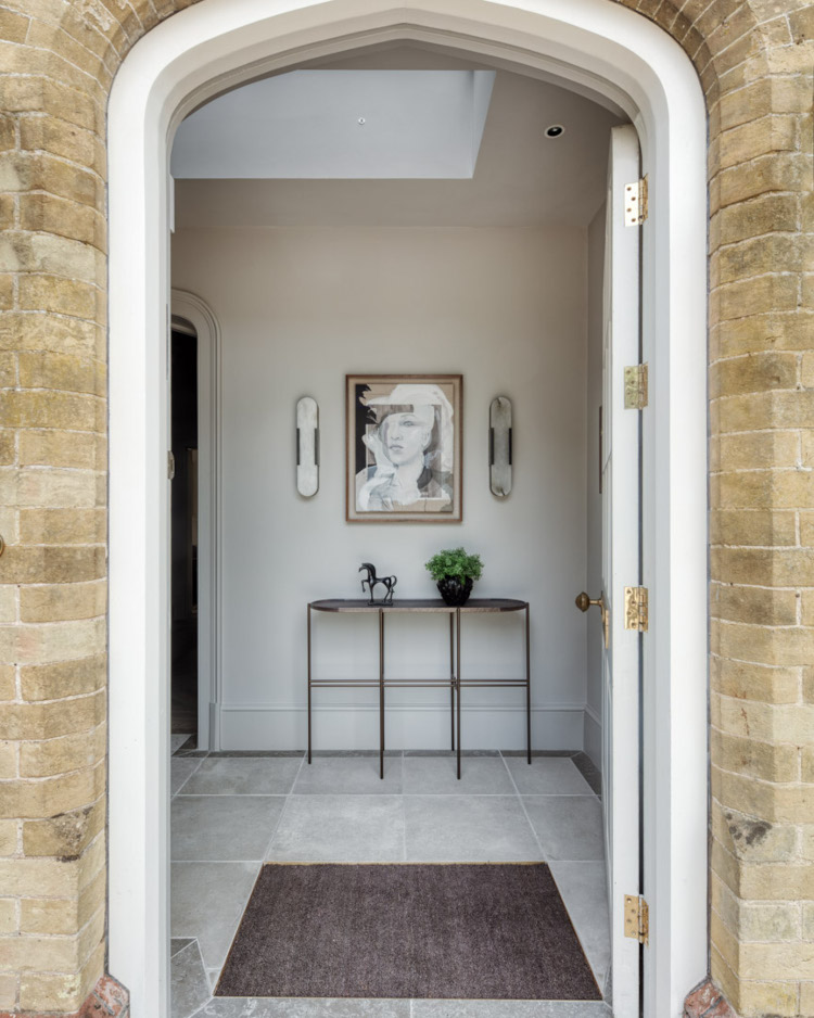 Shot from the outside looking in a large white door is surrounded by honey coloured brickwork. It looks into a elegant polished tile hallway which at the back features a thin console table above which hangs a black and white abstract portrait
