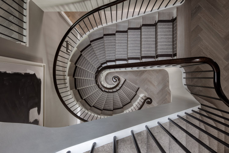Looking from above is the view of a spiral staircase winding down to a hallway. The railings and bannister are made from dark coloured wood and the stairs have a soft grey runner.