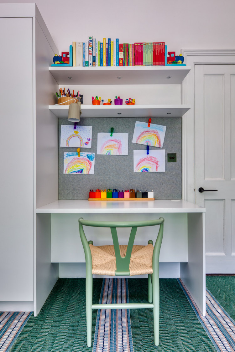 A luxury childrens desk in a playroom featuring a child sized wishbone chair in mint green besides a white wood desk. Behind the desk is a grey fabric pinboard and shelving featuring colourful pictures and toys