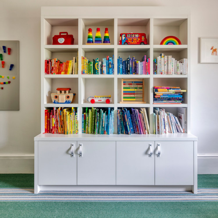 A large white modular bookshelf for a playroom with 16 open shelves featuring a variety of colourful books and toys and 2 white cupboards underneath