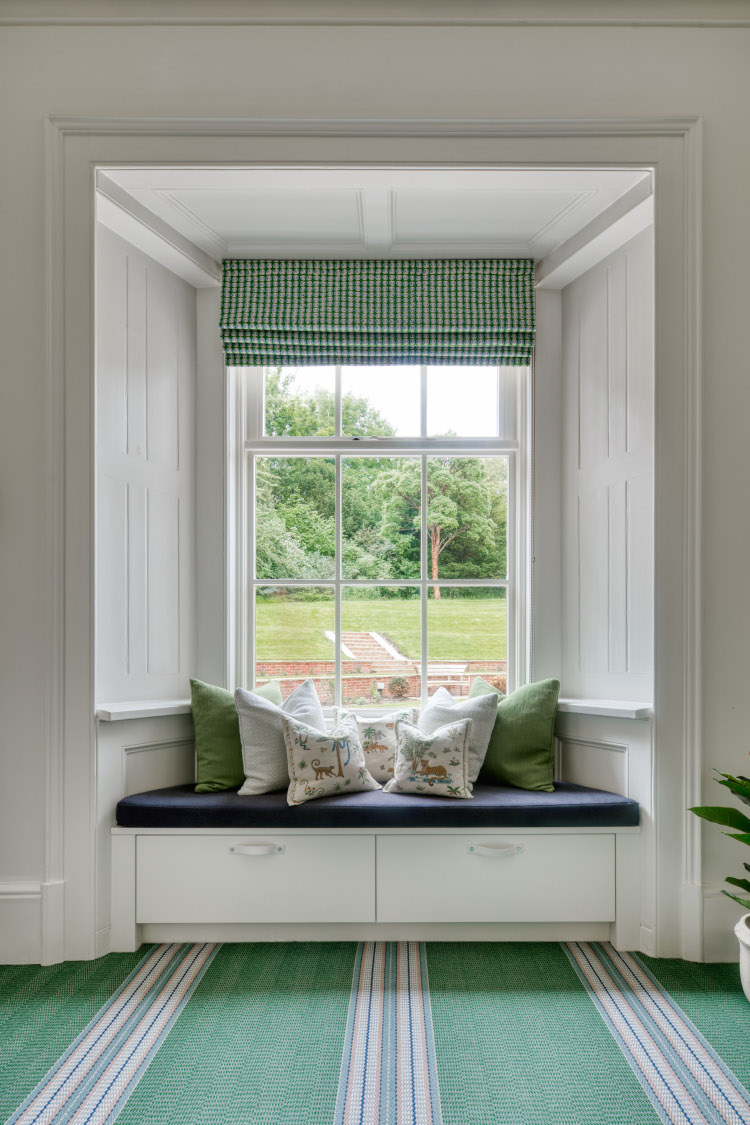 A luxury window seat in a high ceiling Victorian property besides a white sash window with green and white cushions on a navy blue cushion base