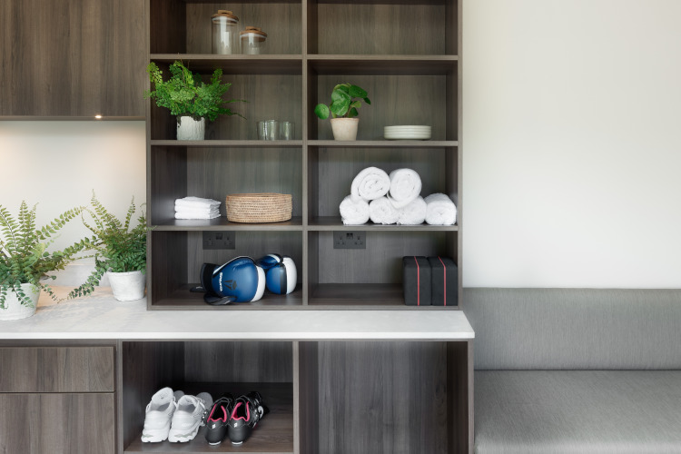 Luxury home gym changing area/storage space showing dark coloured wood shelves and cupboards surrounded by a white topped counter and grey seating area. On the shelves are several white towels, a pair of blue boxing gloves, plants and 2 pairs of exercise trainers.