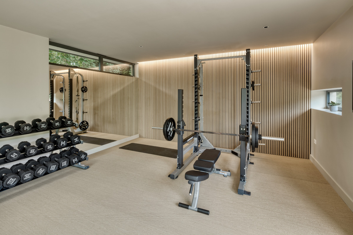 A luxury freeweights area in a home gym showing a loaded barbell bench press, 3 racks of dumbbells with a squat rack behind. Behind is a wood clad wall with hidden lighting coming from above to give the room a large and airy feel.