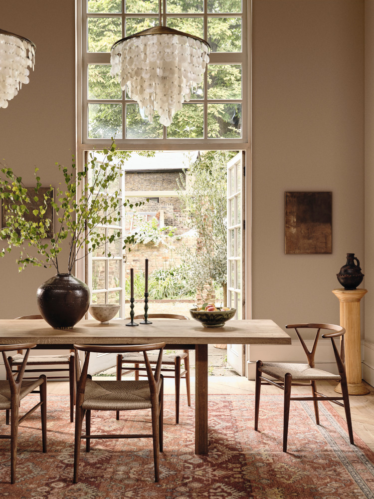 Lick's Beige 02 Paint has been used to paint the Soho Farmhouse which presents an elegeant lightfilled dining room, with 2 chandaliers hanging over a large wooden table, several dark wood wishbone chairs and a red patterned rug. The double french doors behind open out onto a rustic courtyard.