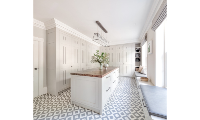 A large white room with patterned tiled floor is a luxury boot room. Around the side are bespoke cupboards in a soft stone colour. In the centre is a freestanding island with draws and to the right are two large window seats with natural light pouring in through the windows.
