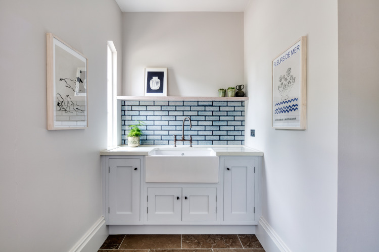 A luxurious Belfast sink with traditional curved tap stands in the middle of the a bootroom flanked on either side by white walls and modern prints in light pine frames. Above the sink is a shelf and a light blue glazed brick splashback with black grouting.