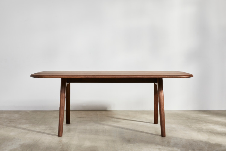 A large brown hardwood dining table from Benchmark Furniture sits in the centre of a bare room with white walls and a wooden floor. A dappled sunlight fills the room.