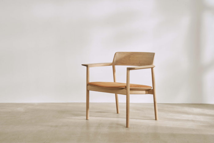 A beautiful light pine chair from Benchmark Furniture sits in the centre of a bare room with white walls and a wooden floor. The chair angled slightly away from the camera and a dappled sunlight fills the room.