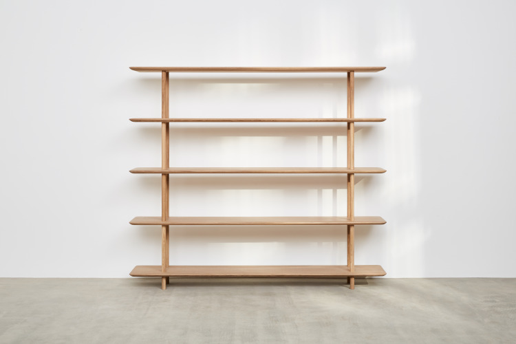 A beautiful light 5 shelf freestanding bookcase from Benchmark Furniture sits in the centre of a bare room with white walls and a wooden floor. A dappled sunlight fills the room.