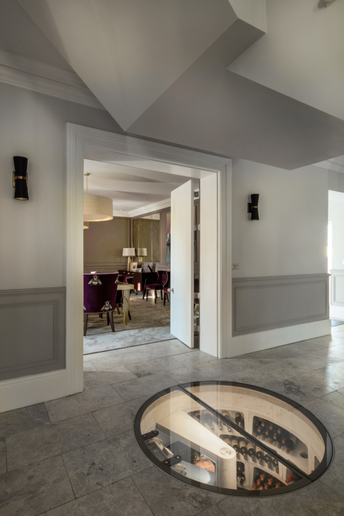 A stunning luxury modern art deco inspired hallway shows the view looking into the dining room. Sitting in a polished grey tile tile floor is a large circular underground wine cellar which is lit showing various bottles of wine.