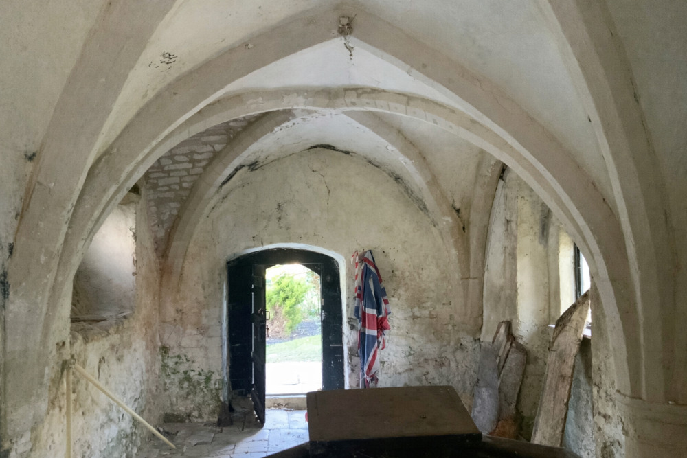 A disused used which will become a guest bedroom in the 15th Century Manor interior design project. A low vaulted ceiling looks out onto a small historic wooden door. Next to it is an old Union Jack flag.