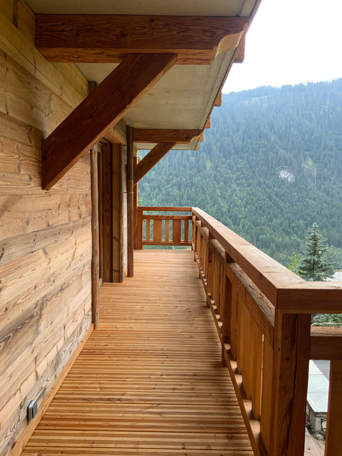 A view of the external walkway / balcony of the Avoriaz chalet with thick, locally cut wooden walkways, wooden walls and wood rails. The view looks out onto a deep, rich Alpine foothills.