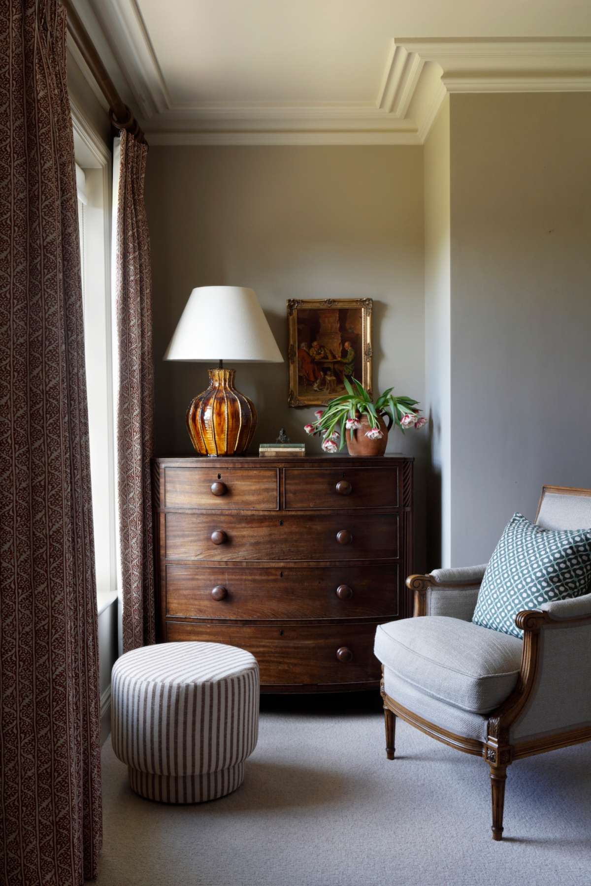 A antique bow front chest of drawers hold a large ceramic table lamp and plant which sits in a recess of the room painted in a soft brown colour. A comfy armchair looks out onto a large bright window which is surrounded by warm floor to ceiling curtains in the West Sussex House interior design project