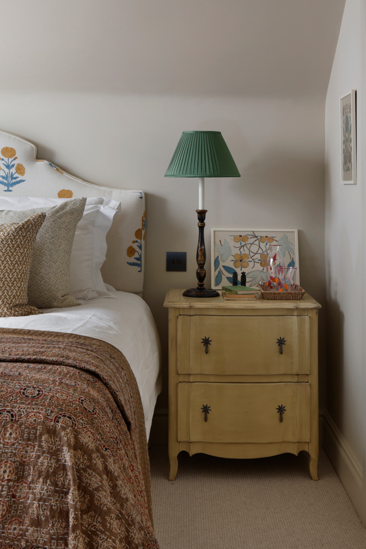 A side view of a guest bed showing floral headboard, a richly coloured patterned quilt, soft wood bedside table, bedside lamp as part of the West Sussex House interior design project.