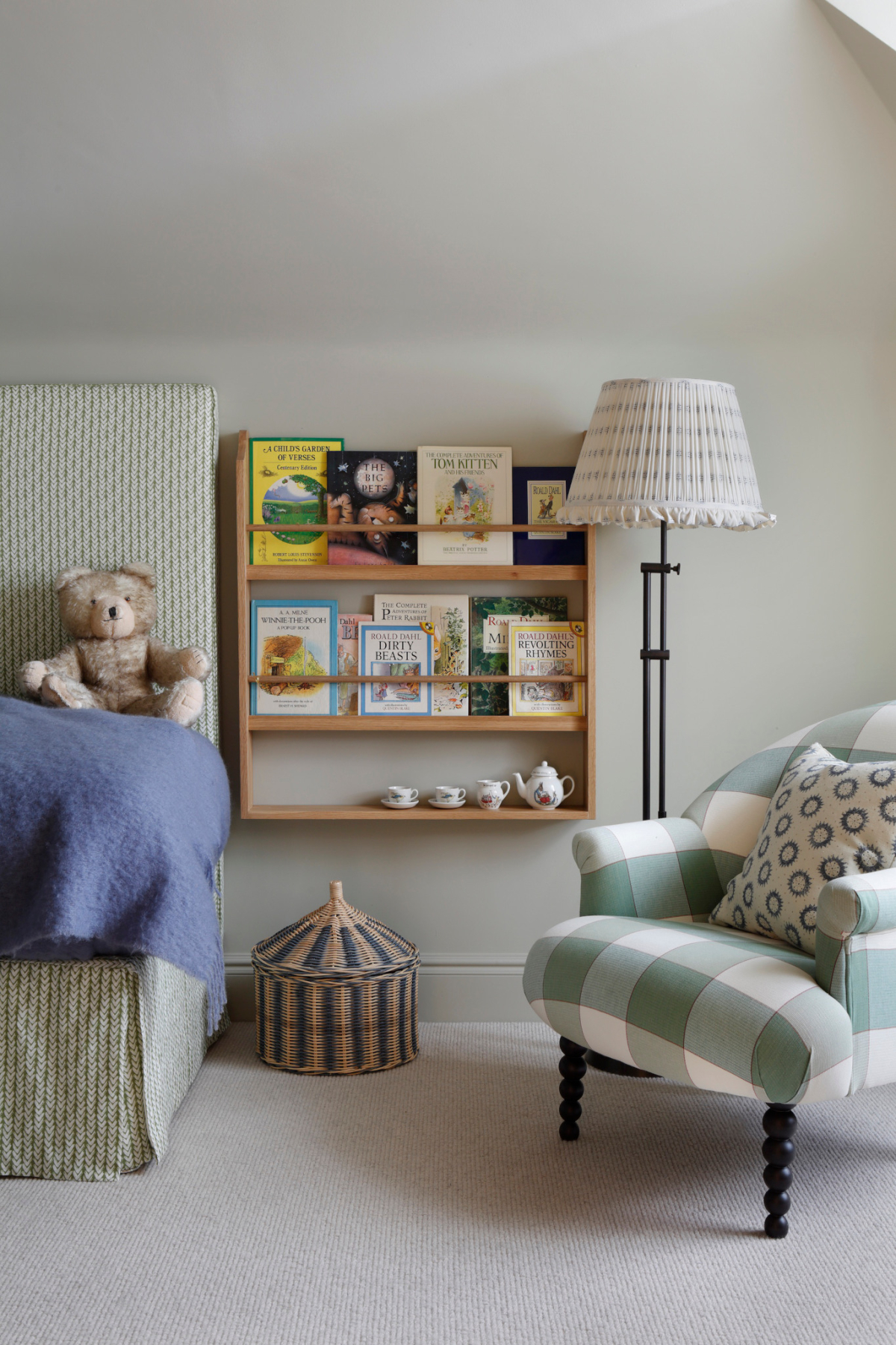 A side view of the childrens bedroom in the West Sussex House interior design project showing bed with teddy bear, small armchair in a green gingham, book shelf, floor lamp, and basket. The carpet is a soft beige and the walls a soft sage colour.