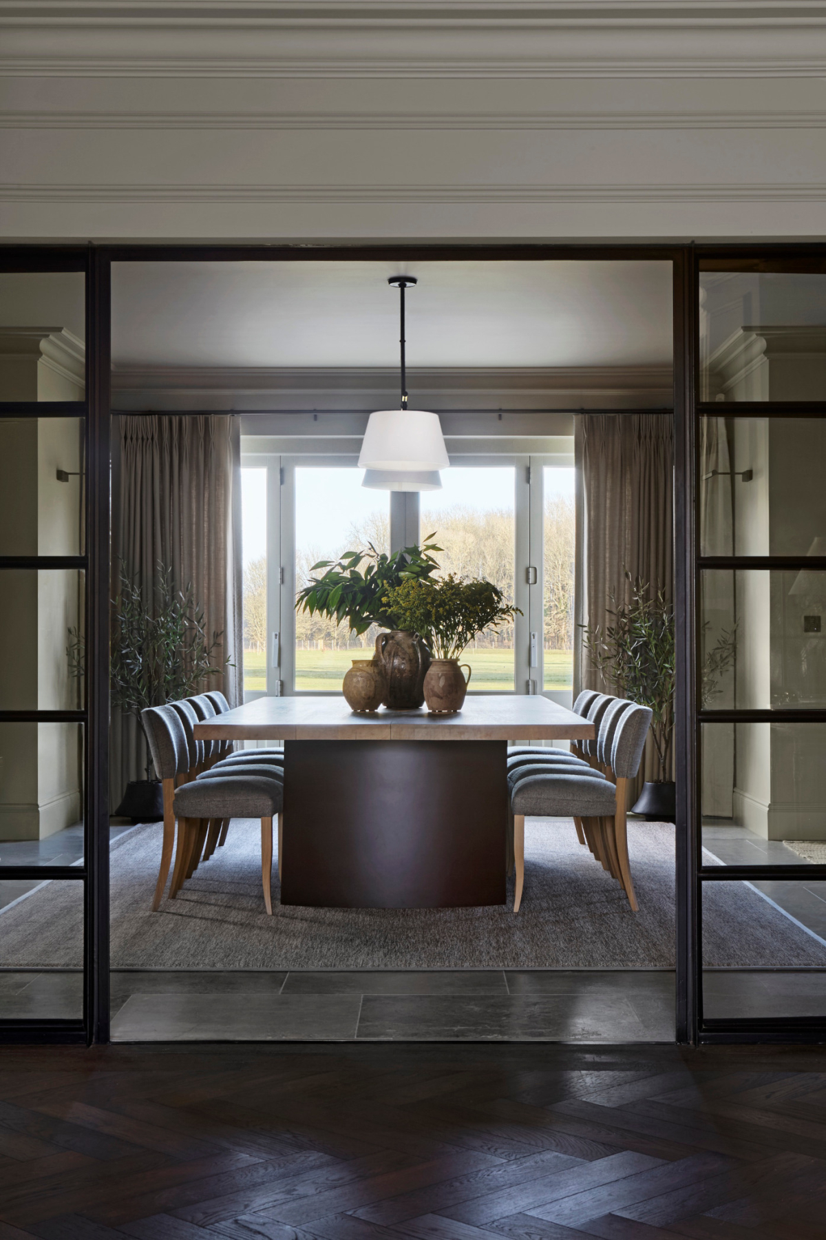 A large modern dining room in the West Sussex House interior design project with dining table and chairs frames the picture which is fronted by modern glass panelled sliding doors and a wooden floor. The room is illuminated by a strong white light coming from windows at the rear with views out onto the surrounding gardens.