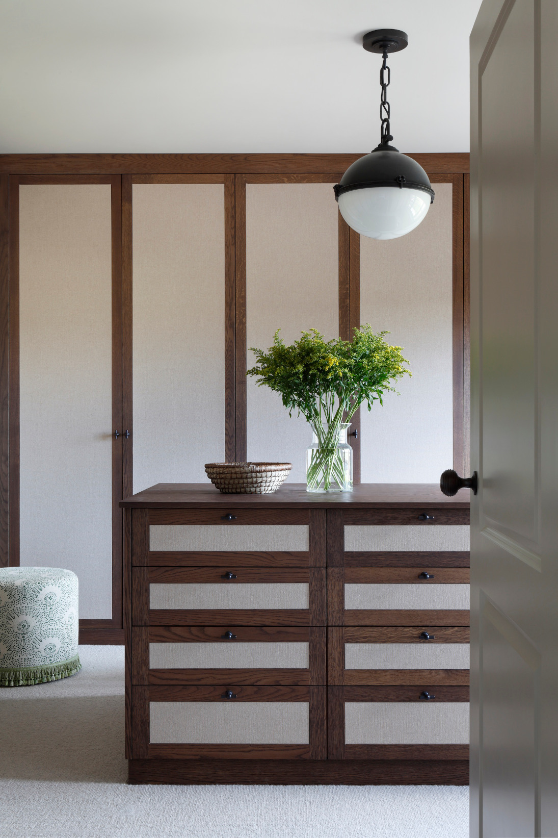 Taken from the doorway the camera looks onto a luxury dressing room of the West Sussex House interior design project complete with dark wood floor to ceiling cupboards and drawers. From the roof a large black metal lampshade hangs with a frosted white bulb..