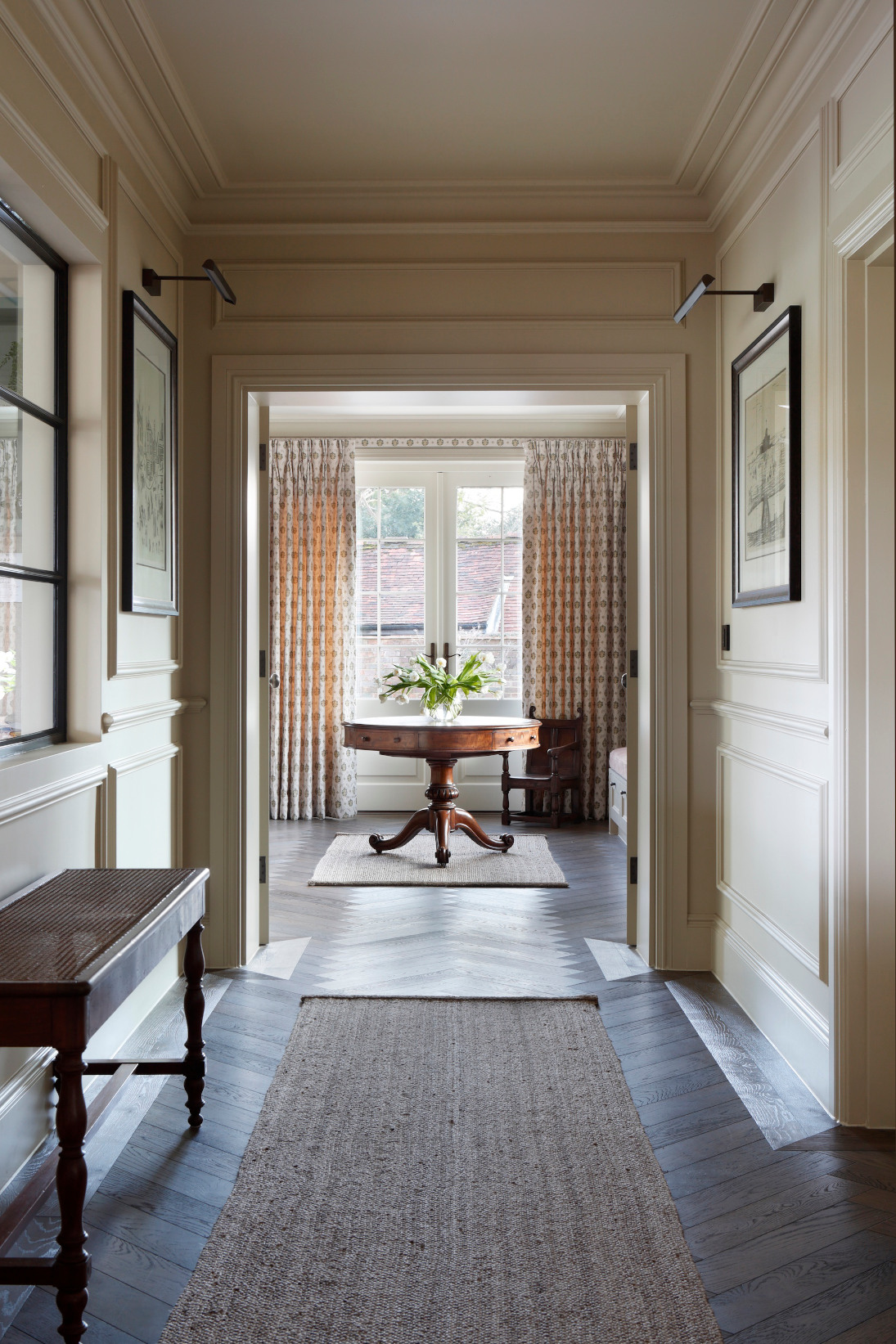 A long entrance hallway showing various pictures on the walls centres on a large, circular hardwood table which is framed by a large window and floor to ceiling patterned curtains. Part of the West Sussex House interior design project.