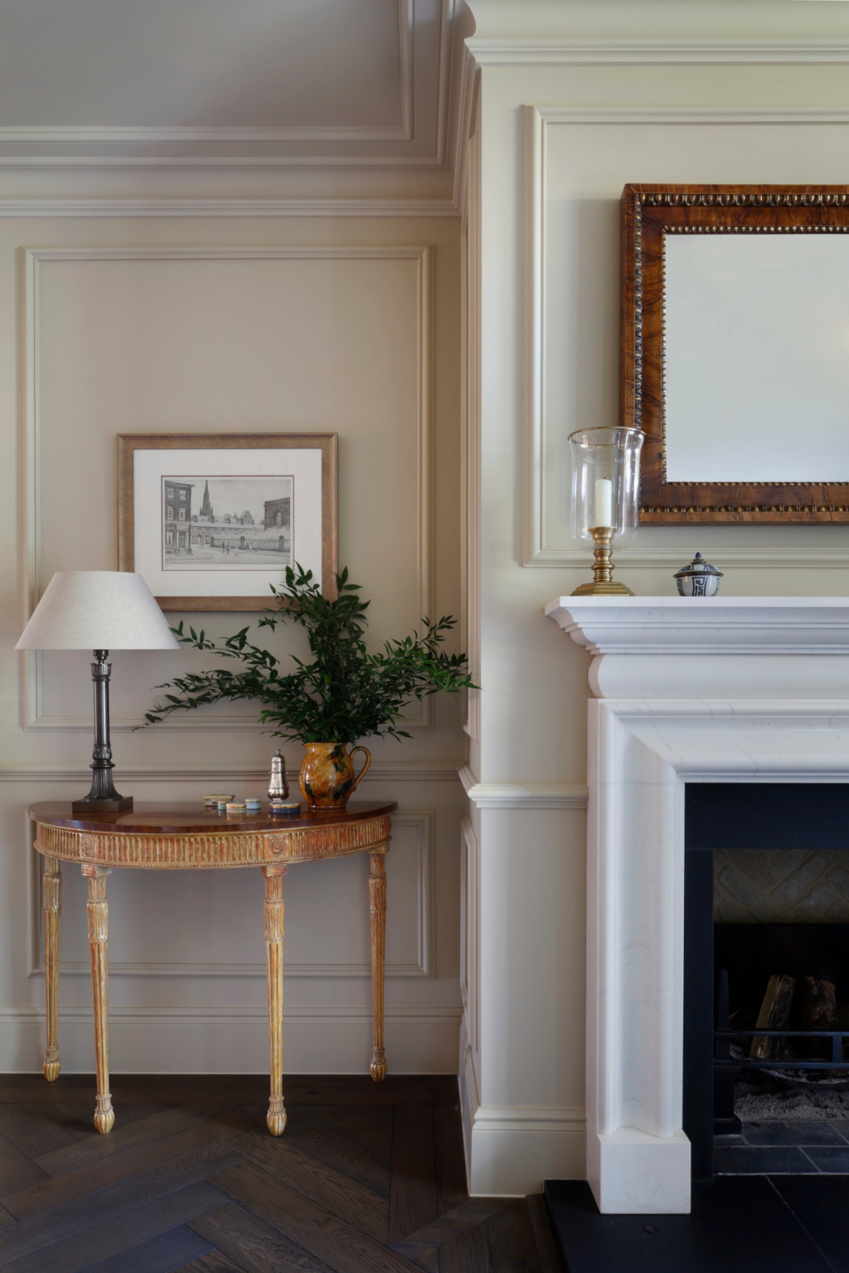 The side view of a large marble fireplace surround in the West Sussex House interior design project. to the left is an antique console table on which sits a table lamp and plant. The panelled walls are a soft yellow colour.