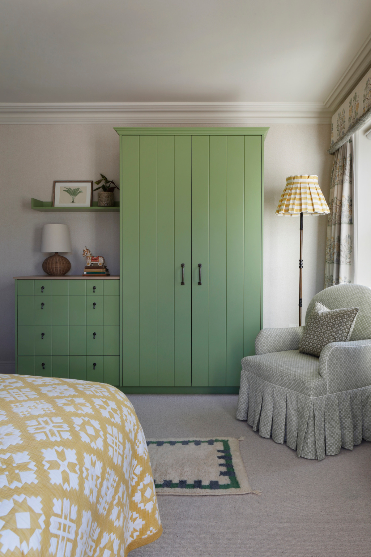 A large mint green cupboard and chest of drawers frame the guest bedroom of the West Sussex House interior design project. To the foreground is a grey coloured armchair and yellow and white patterned quilt on a double bed.