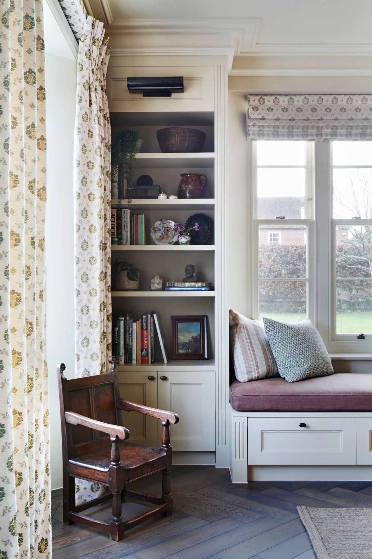 The library and reading room of the West Sussex House interior design project. A small antique wooden chair sits besides large patterned floor to ceiling curtains and a bookshelf. To the right is a plum coloured padded window seat with views out onto the garden.