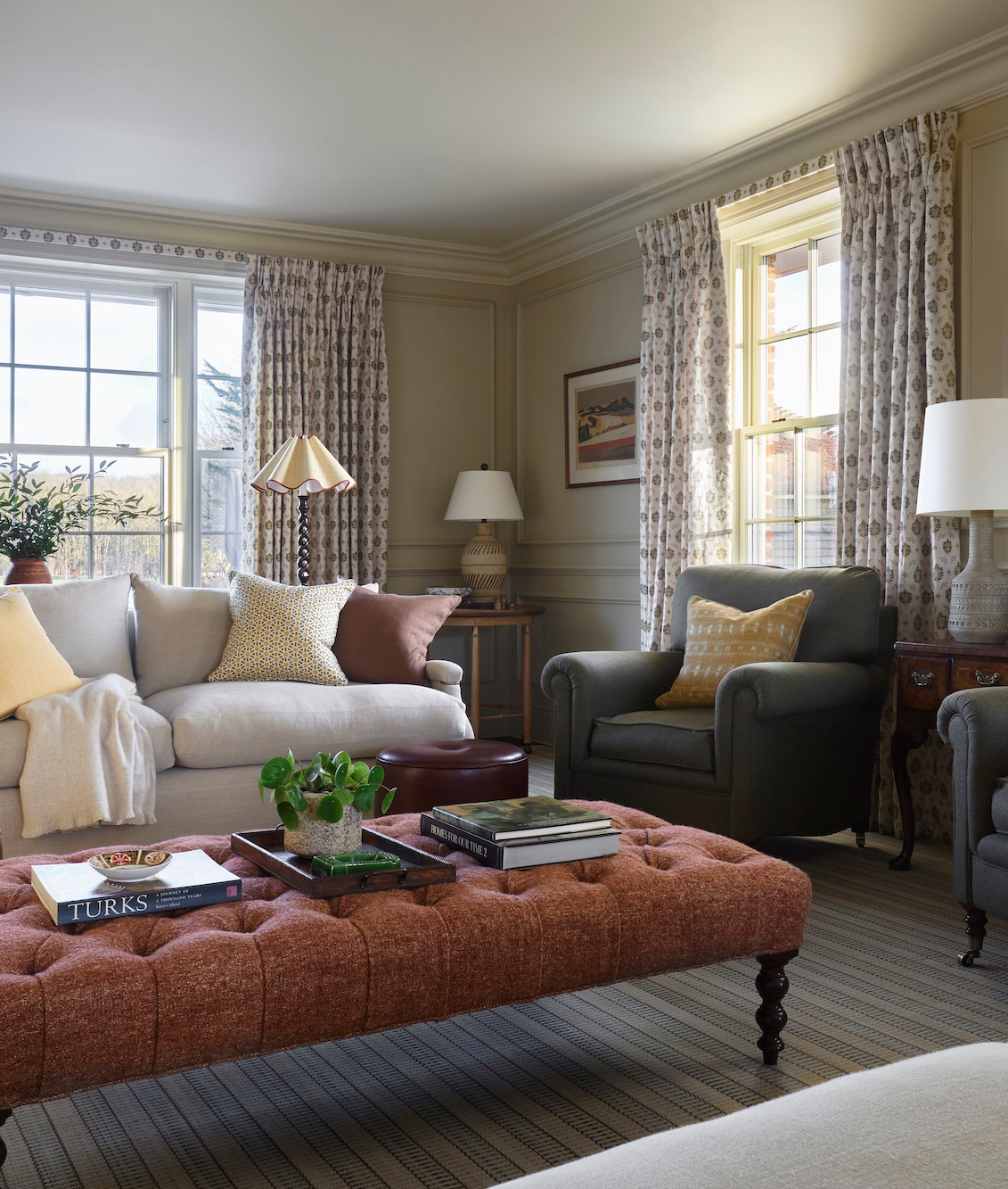 A large comfy living room that is part of the West Sussex House interior design project. A armchair and sofa are full of cushions while floor to ceiling patterned curtains contain large french windows. To the front is a large terracotta red pouffe on which several books and magazines sit.