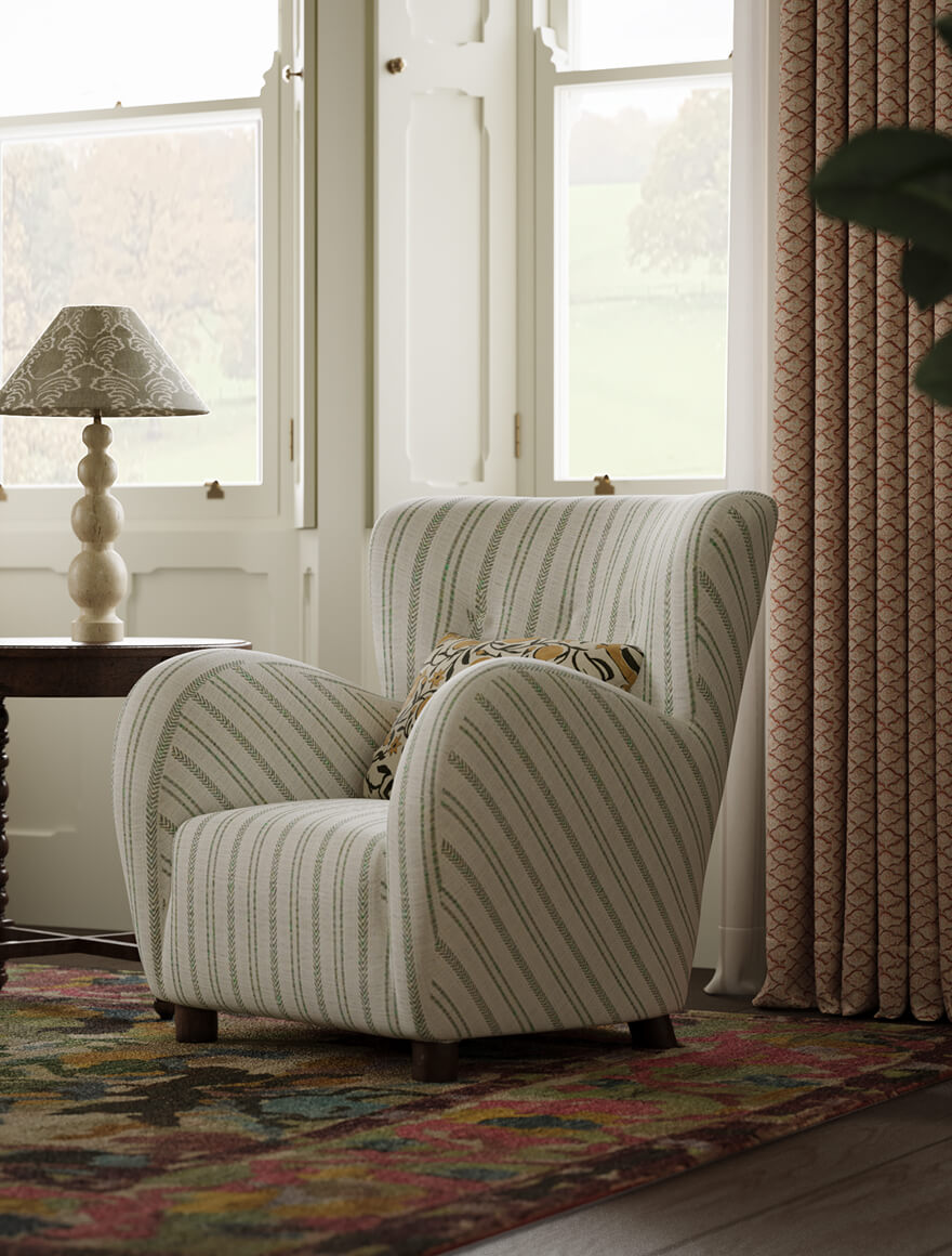 A large a comfy white upholstered bedroom armchair sits in a stunning English country property bedroom with textured rug, bedside lamp. Behind are red patterned floor to ceiling curtains and white shuttered windows looking out over the English countryside beyond.