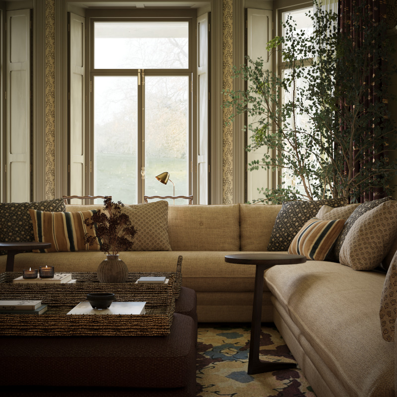 A view of the living room of the Queen Anne manor interior design project. Taken from a low view it looks out over a coffee table and large neutral coloured wraparound sofa to a large house plant and shuttered windows which look out onto the English countryside beyond.