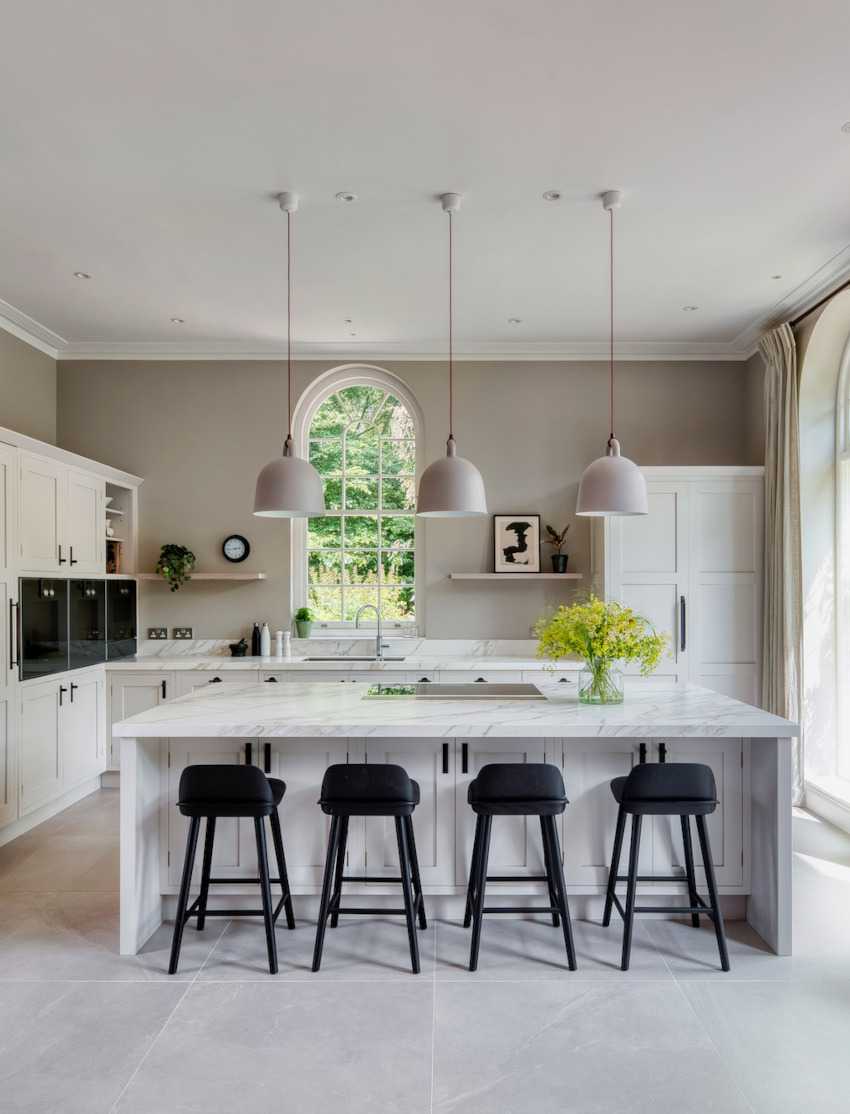 A bright modern luxury kitchen with large central kitchen island and 4 black stools occupy the center of the room. Around the sides are various cabinets and suspended from the ceiling are 3 long taupe coloured metal lampshades. Used for the kitchen design image for Studio Hooton's interior design services.