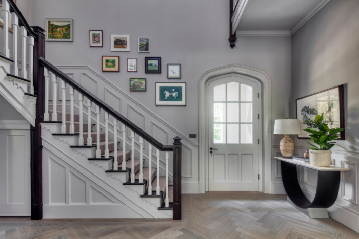 A large hallway showing a herringbone floor, staricase with assorted pictures running up the side of it while to the right stands a large white traditional front door and entrance table