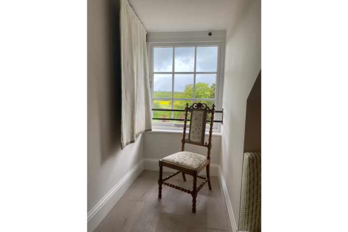 A historic brown wood flat backed chair with upholstered seat and barley twist legs sits in a small recess of a historic property besides a square sash window. Outside you can see various green fields and trees.