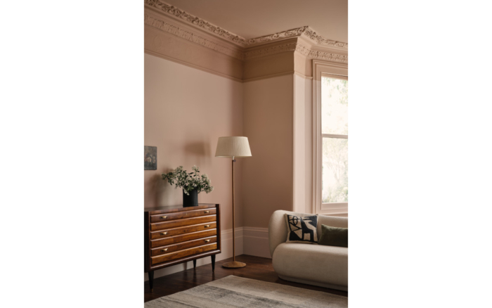 A high ceilinged living room with ornate coving sets the backdrop to dusted rose coloured walls, a dark brown wooden dresser, white sofa, white floor lamp and large light colour floor rug
