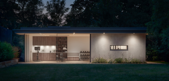 A photo taken at sunset looking out from a garden towards a custom built building housing a luxurious home gym. Spotlights from the roof casts shadows onto a changing area where in front stands an exercise bike, rowing machine and dumbbell rack.