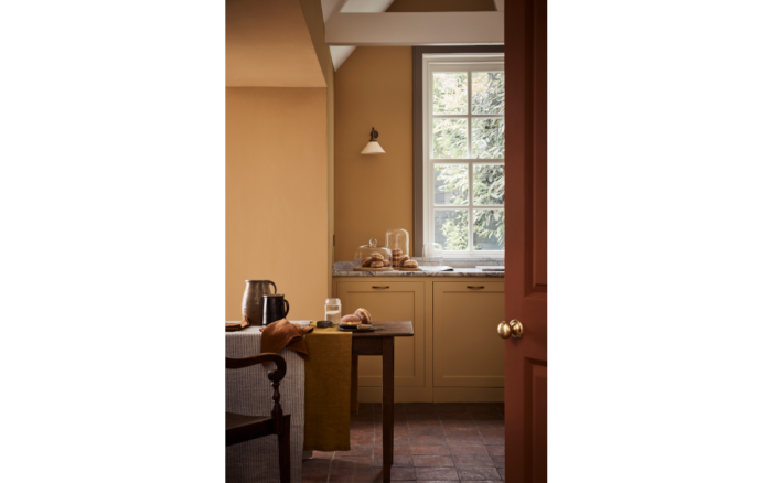 A rustic kitchen is painted using Little Green's Bombolone paint. A dark wooden table holds several glasses next to an anytique dining chair. Towards the back of the room are two cupboard and countertop which sit below a sash window.