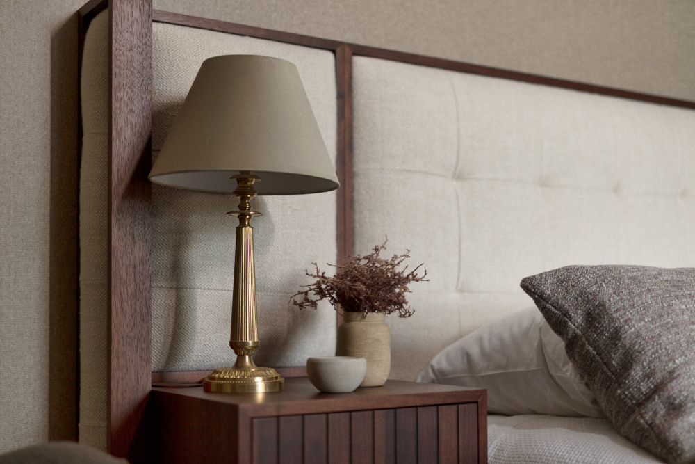 A close up image of a quilted beige headboard with a hardwood surround. To the left is a hardwood bedside table with a small plant and a large muted copper coloured beside lamp with a stone coloured lampshade.