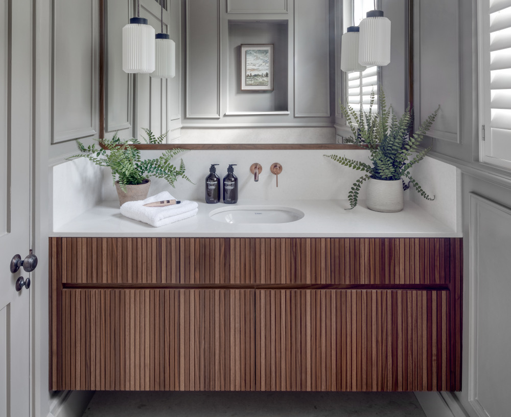 A beautiful dark wood panelled vanity unit sits as the centre of the cloaroom. A single basin is sunk into a marble countertop and begind is a full length mirror with two white hanging ceiling lights. The reflection shows the luxurious grey wood panelling of the wider room.