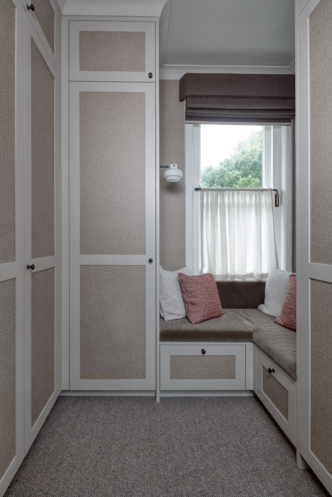 A luxurious dressing room made of custom cabinets in earth colours is seperated with a brown coloured seating area where dusty pink and cream white scatter cushions are positioned. Behind is a small window, half covered with netting which looks out onto the countryside.