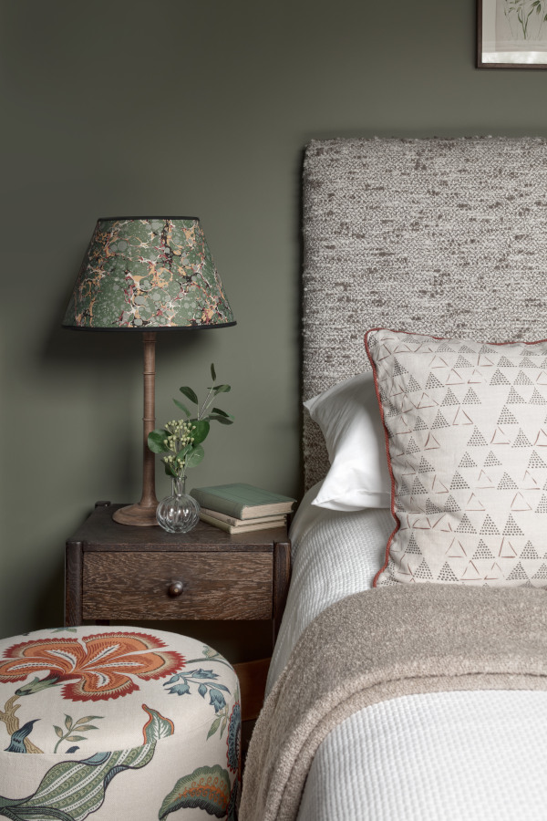 One half of a luxurious double bed in the guest bedroom shows a grey fabric headboard, a floral pattern footstool alongside a dark wood bedside table which has a slender beside lamp with floral lampshade. Behind the walls are painted in a deep olive green.