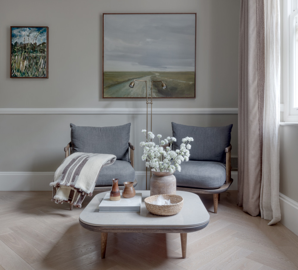 A stunning light coloured herringbone floor is shown on which sits a small square coffee table with rounded corners which sits in front of two beautiful grey armchairs with dark wooden frames. On the table is several pots including a cermaic flowerpot with white flowers. The stone coloured walls feature 2 small original artworks of landscape scenes and to the right are light brown coloured curtains around a window which sends in a strong natural light.