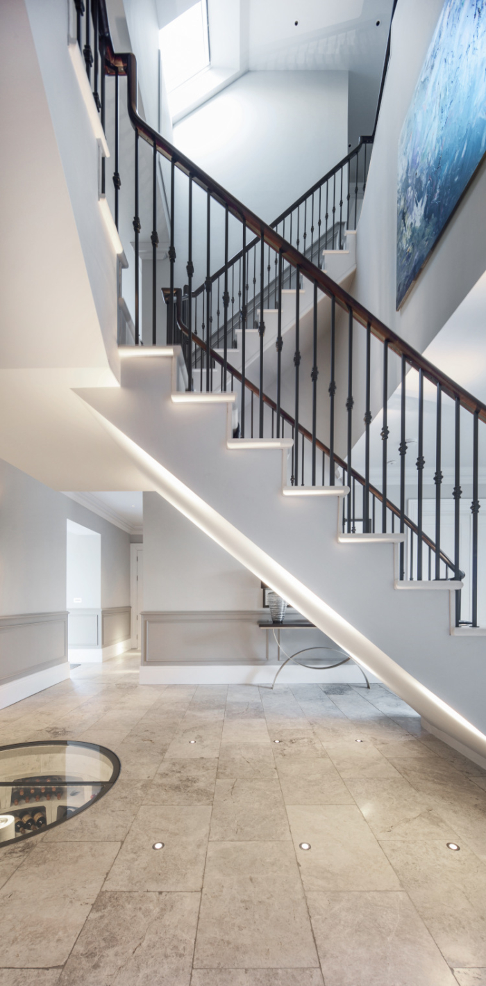 A polished tiled floor shows the steep underside of a modern white staircase which is illuminated by floor lighting.
