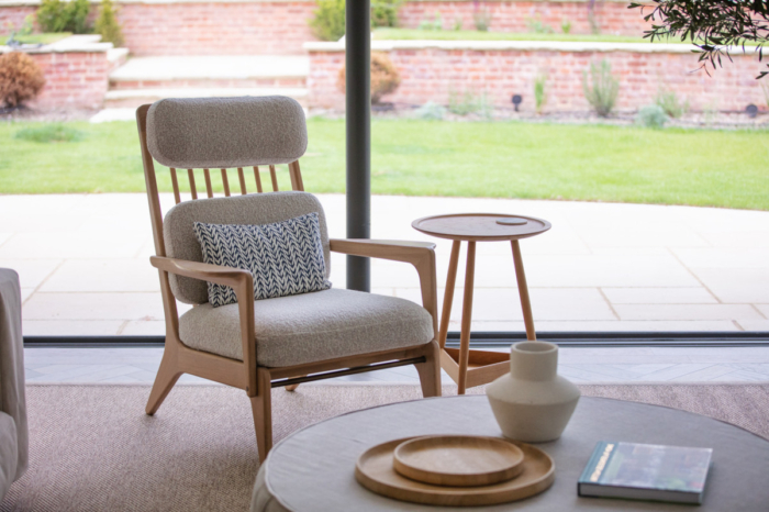 An oak coloured armchair with oak side table made by Benchmark furniture sits in a large modern lounge in front of a huge glass door which you can see through to a large patioed garden.