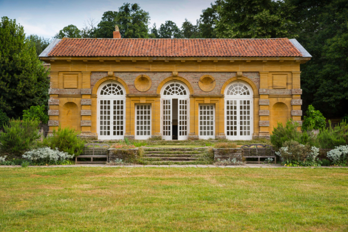 An exterior front view of an orangery. Tall grand full height white surrounded windows and doors featuring the arch top sit across the front facade of the orangery. Steps lead up to the middle set of window doors across the grass.