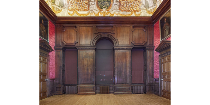 A historic room within a medieval building featuring dark solid wood panelling, ornate decorative ceiling and deep red patterned wallpaper. A large traditional heavy door at either end of the space has a grand framed portrait hung above.