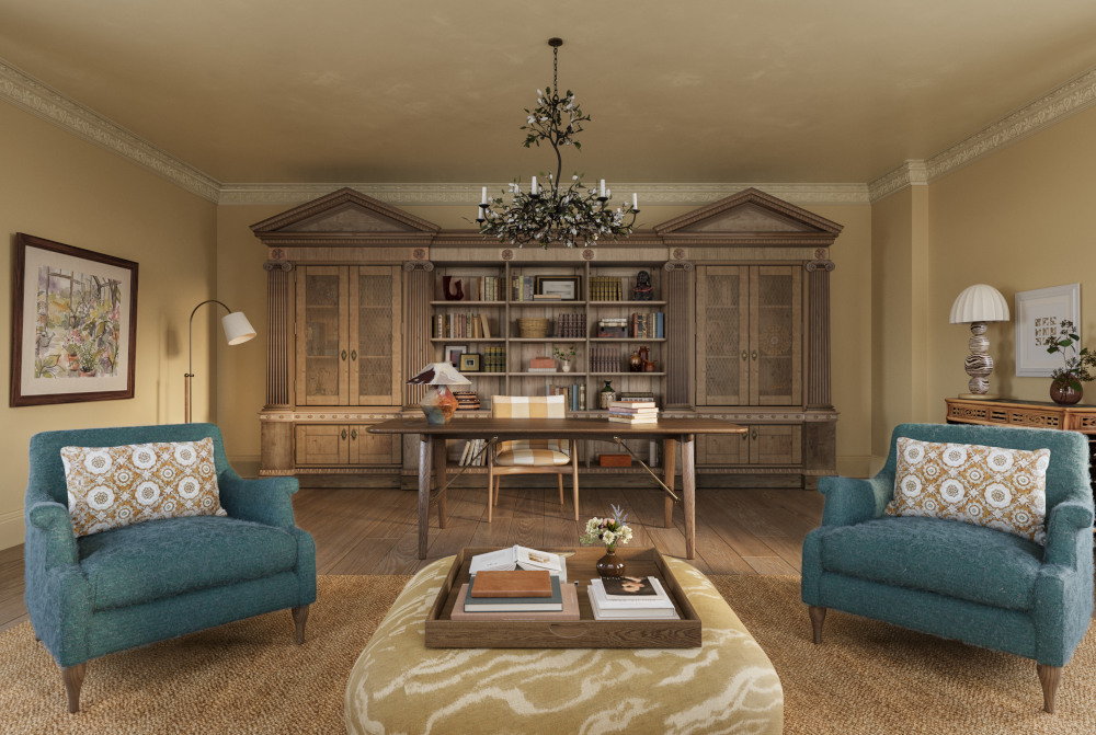 A symmetrical image of the Sussex estate library showing a grand mustard colour room. In the background is a dark wood classical shaped set of shelves and cupboards, a dark wood desk above which a black chandelier is suspended. In the foreground two teal coloured armchairs with floral patterned cushions sit either side of a large mustard coloured pouffe on which sits a tray of books and objects.